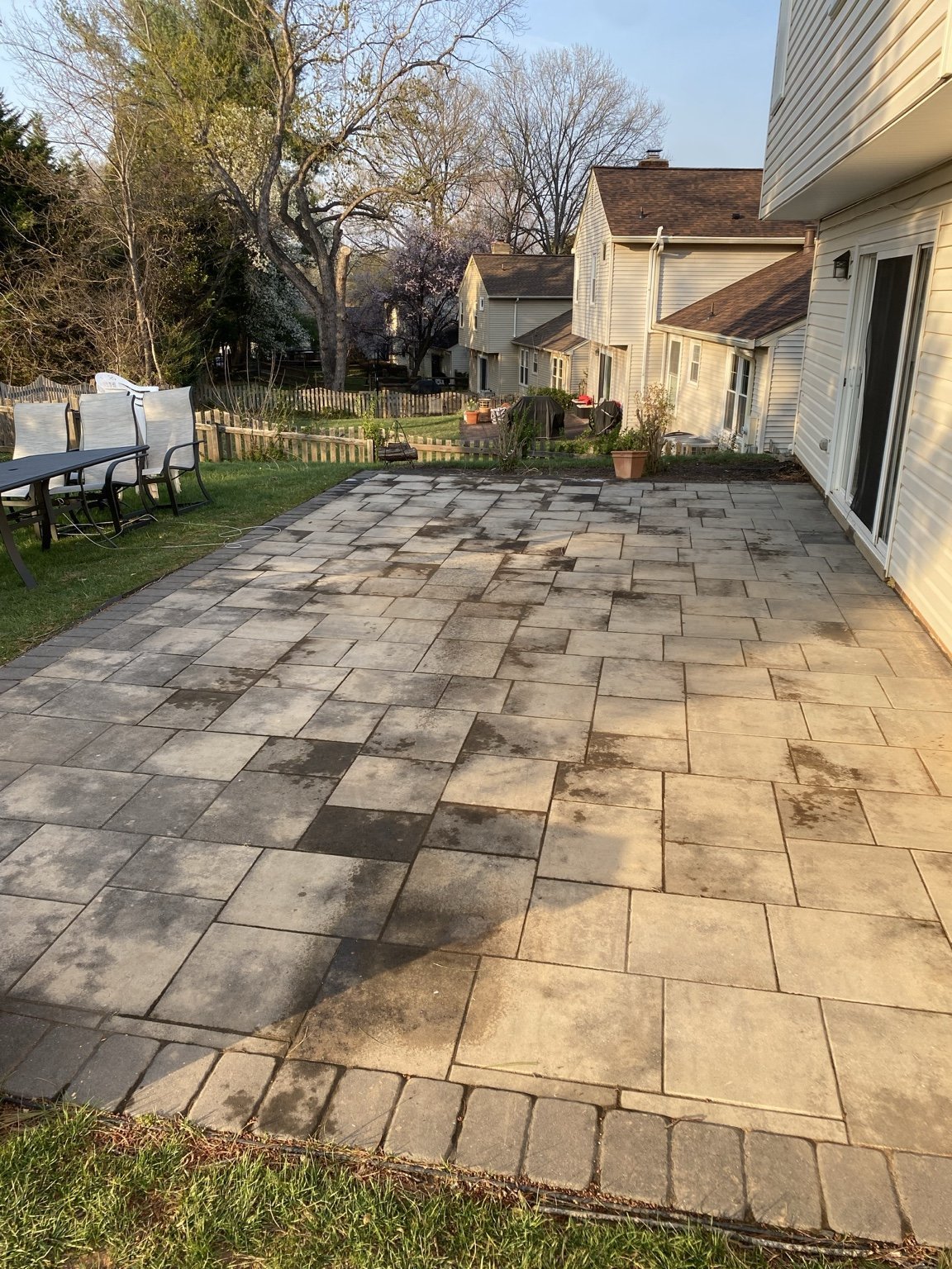 Spacious backyard patio with stone pavers, white chairs on left, fenced garden area, and residential houses in background on clear day