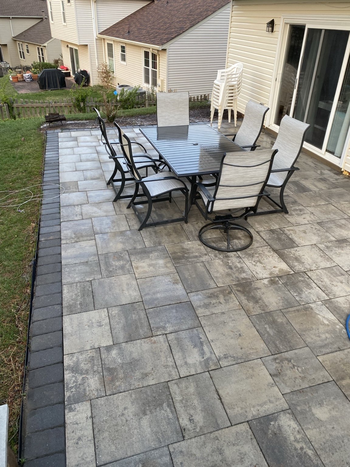 Backyard patio with stone pavers, outdoor dining table with black metal chairs and white cushions, adjoining a residential home with white siding